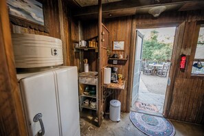 Un interior rústico de madera con un refrigerador blanco, estantes con utensilios de cocina y una puerta que da a un área de estar al aire libre.