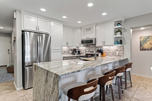 A modern kitchen with a stainless steel refrigerator, a microwave, and a coffee maker on the countertop. There are four bar stools and a large marble island.