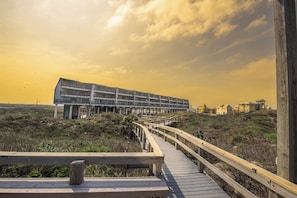 A wooden boardwalk leading to a modern building with multiple balconies, surrounded by natural landscape.