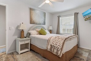 Ein Schlafzimmer mit einem Bett, zwei Nachttischen, einem Deckenventilator, einem Fenster mit Vorhängen und einem an der Wand hängenden Kunstwerk.