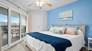 A bedroom with a bed, a ceiling fan, a painting, and a view of the outside.