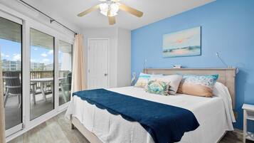 A bedroom with a bed, a ceiling fan, a painting, and a view of the outside.
