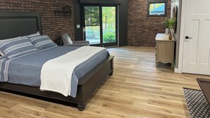 A bedroom with a brick wall, a large bed with a headboard, a wooden floor, and a sliding glass door leading to a balcony.