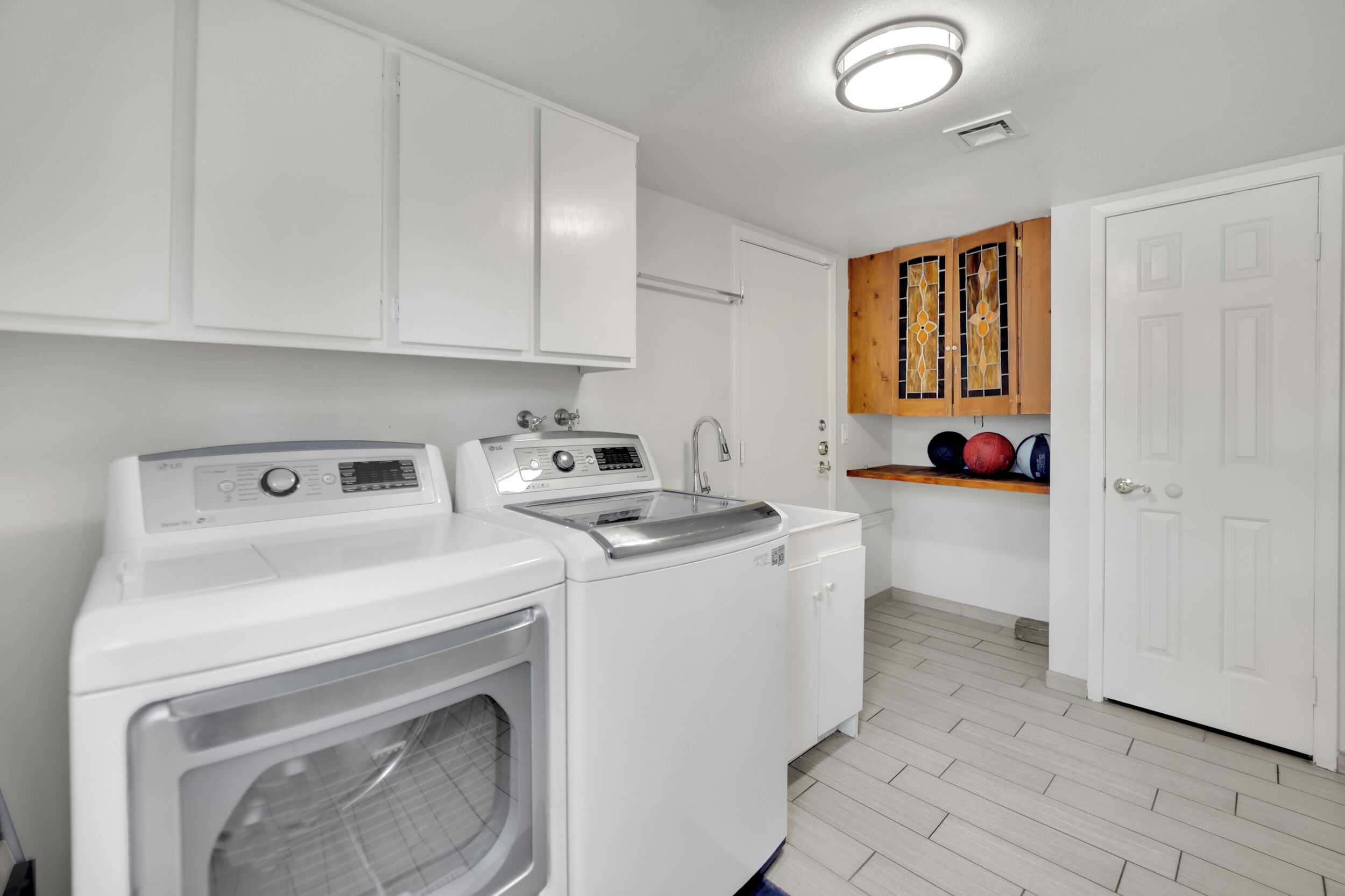A laundry area with two washing machines, a dryer, and a wooden shelf with sports balls.