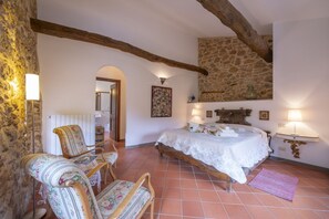 A bedroom with a stone wall, wooden beams, a bed, two chairs, and a nightstand with a lamp.