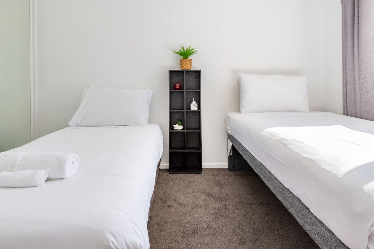 Een hotelkamer met twee eenpersoonsbedden, een klein zwart plankje met een plant erop, en een witte handdoek op het linkerbed.