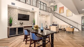 A spacious living area with a herringbone wood floor, a dark wooden dining table with striped chairs, a flat-screen TV mounted on a stone wall, a staircase with a metal railing, and a large window providing natural light. - Stay longer, Spend Less, 2 Master Suites! The Cays (Chandler)