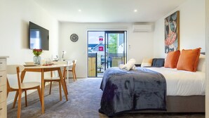 Une chambre moderne avec un lit, un bureau et une chaise. Un téléviseur est fixé au mur et une fenêtre offre une vue sur un bâtiment à l’extérieur.