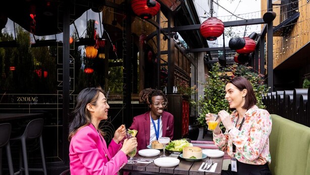 Un groupe de personnes est assis à la terrasse d’un restaurant dont l’architecture moderne est remarquable, avec des lanternes rouges et une enseigne bien visible sur laquelle on peut lire « ZODIAC ».