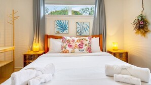 Una cama bien hecha con almohadones florales, dos lámparas de noche y vistas al exterior con vegetación.