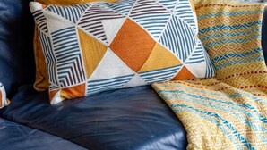 Un sofá de cuero azul con cojines estampados, una escalera blanca y una estantería con libros y objetos decorativos.