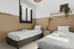 Ein Hotelzimmer mit zwei Betten, einem Fenster mit Jalousien, einem Nachttisch mit Lampe und einem an der Wand befestigten Regal mit Pflanze.