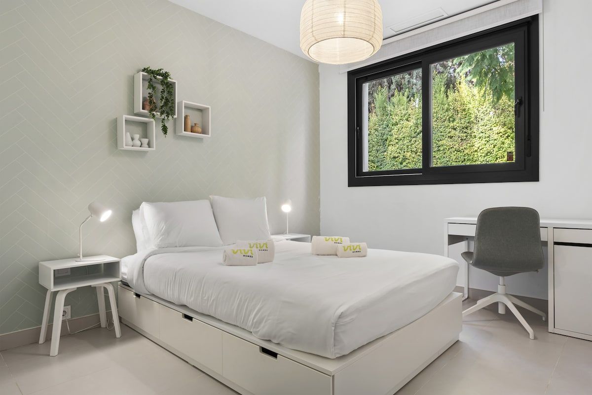 A modern bedroom with a bed, bedside tables, a desk, and a window with a view of greenery.