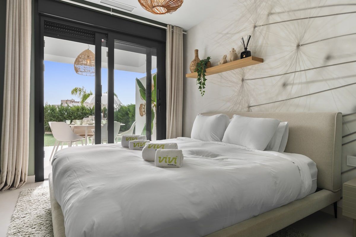 A modern hotel room with a large bed, a sliding glass door leading to an outdoor area, and a small shelf with decorative items.