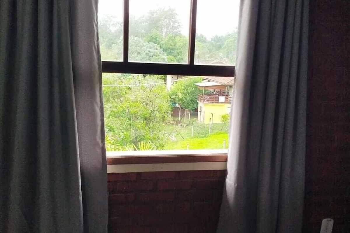 Um quarto com uma cama, uma janela com cortinas e vista para o verde lá fora.