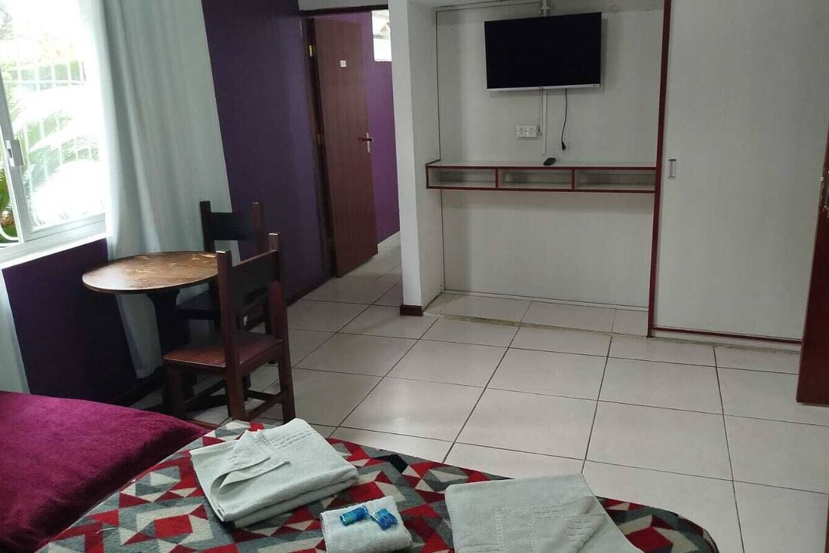 Um quarto de hotel pequeno com uma cama, uma cadeira, uma mesa, uma televisão e uma porta.