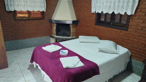 A room with a bed, a fireplace, and a window with lace curtains. - Pousada Vale das Flores (Itatiaia)