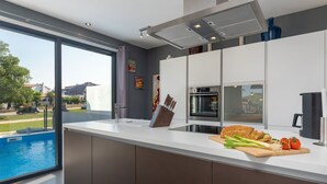 A modern kitchen with a large island, a built-in oven, and a view of a swimming pool. - Villa Maestral (Istria - Funtana) (Funtana)