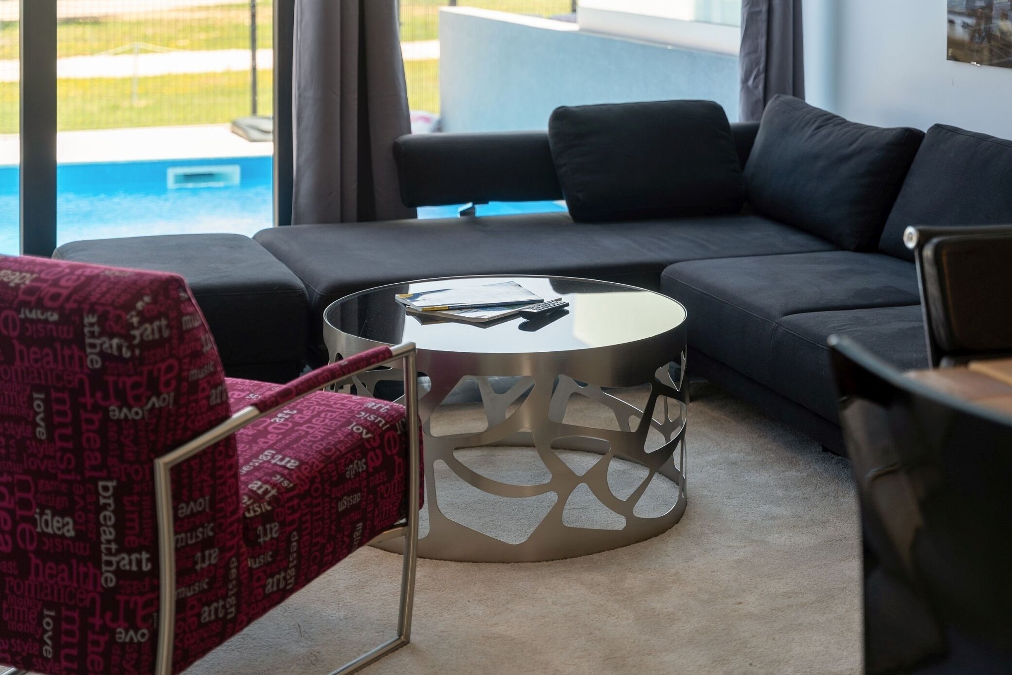 A modern living room with a black sofa, a round glass coffee table, and a red patterned chair.