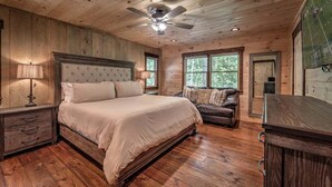 A bedroom with a large bed, a wooden headboard, a ceiling fan, and a window with a view of trees. - Dialed Inn- Pet Friendly | Dock on Private Lake | Arcade Games (Morganton)