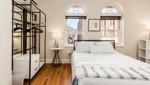 A bedroom with a bed, a wardrobe, a round side table, and two windows with arched tops.
