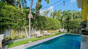 A swimming pool with a clear blue water, surrounded by lush greenery and palm trees. There are two lounge chairs and a hammock. - 1009 Managed by Brampton Park (Delray Beach)