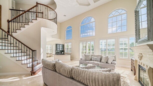 A spacious living room with a large gray sofa, a glass coffee table, and a fireplace. A spiral staircase with black railings leads to an upper level. The room has large windows with arched tops, allowing natural light to fill the space.