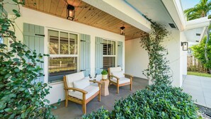 A covered porch with two wooden chairs and a small table, surrounded by lush greenery. - 246 Managed by Brampton Park (Delray Beach)