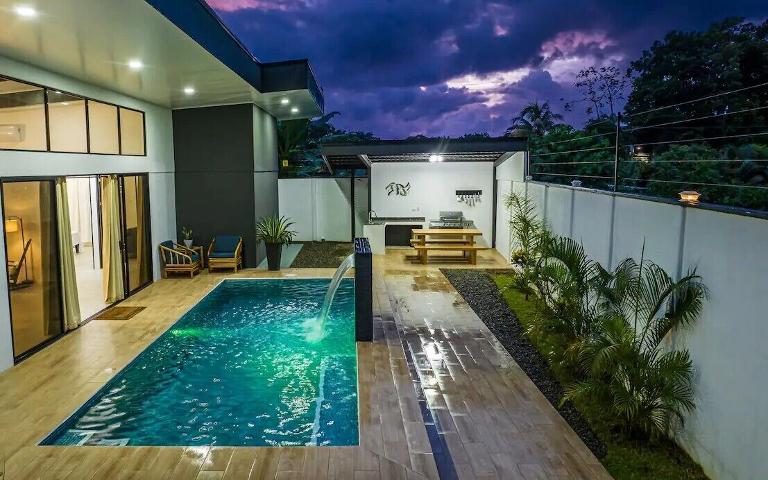 Un área de piscina exterior moderna con terraza de madera, una zona de barbacoa y vista al cielo.