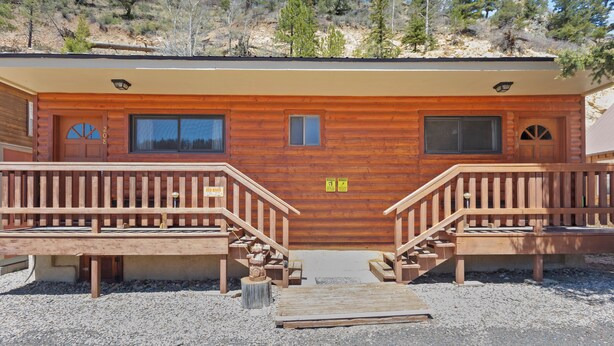 Une cabane en rondins avec une terrasse en bois, deux escaliers et une allée gravillonnée.