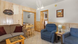 Un salon chaleureux avec un mur en bois, un fauteuil bleu, une petite table et un coin repas avec des tabourets.