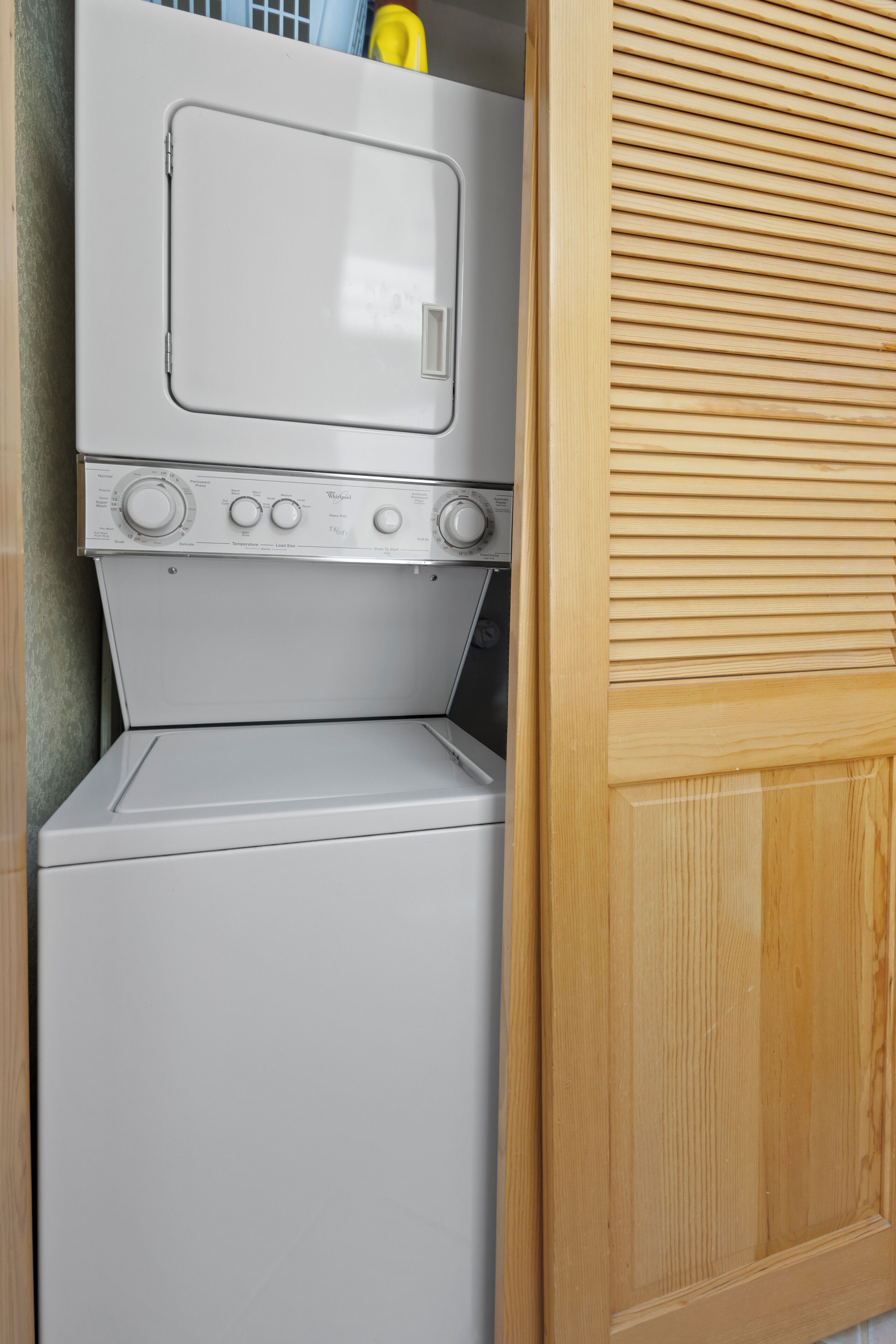 A compact laundry room with a front-loading washer and dryer, wooden blinds, and a small shelf with a yellow item.