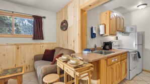 Une cuisine compacte avec des meubles en bois, un coin repas avec des tabourets, un canapé et une fenêtre donnant sur la forêt.