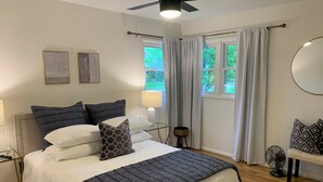 Un dormitorio con cama, ventilador de techo, ventana con cortinas y mesita de noche con lámpara.