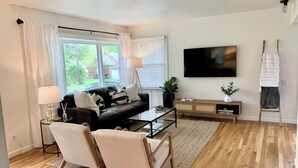 Una sala de estar moderna con un sofá de cuero negro, dos sillones beige, una mesa de centro de vidrio y un mueble de televisión de madera.