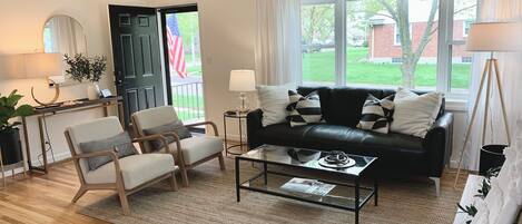 A living room with a black leather sofa, two armchairs, a glass coffee table, and a large window with curtains.