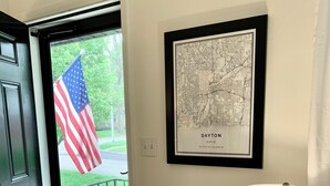 Un pasillo con un mapa enmarcado de Dayton, Ohio, una puerta con una bandera estadounidense, una mesa de vidrio con una lámpara y un par de zapatos sobre una alfombra.