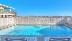 A clear blue pool with white lounge chairs in the foreground and a wooden deck with a railing in the background.