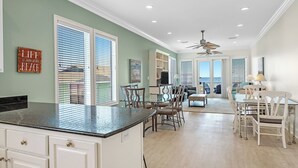 A spacious kitchen with a large island, dining area with white chairs, and a view of the ocean through the sliding glass doors.