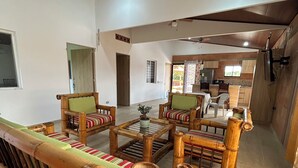 A spacious living area with a wooden sofa set, a dining table, and a kitchen in the background.