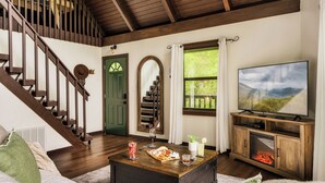 A living room with a wooden staircase, a fireplace, a television, and a dining area with a table set for two. - Cozy Getaway From It All ~ Heated Pool ~ Sauna ~ Hot Tub ~ Games ~ A-Frame (Sevierville)