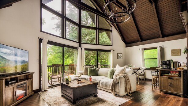 A spacious living room with a large TV, wooden furniture, a fireplace, and large windows offering a view of the outdoors. - Cozy Getaway From It All ~ Heated Pool ~ Sauna ~ Hot Tub ~ Games ~ A-Frame (Sevierville)