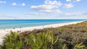 Een strand met helderblauw water, een zandstrand en schaarse begroeiing op de voorgrond.