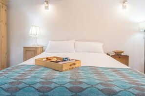 Una cama bien hecha con una colcha azul estampada, una mesita de noche de madera con una lámpara y una bandeja con desayuno sobre la cama.