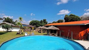 A swimming pool with a wooden deck, surrounded by a fenced area with a red building and thatched-roof structures.