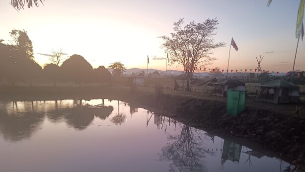 A calm body of water reflecting trees and a clear sky. There are tents and flags visible, suggesting a campsite or outdoor event setting.