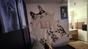 A bedroom with a cowhide wall art piece labeled "RODEO," a bed with multiple pillows, and a large framed picture on the left.
