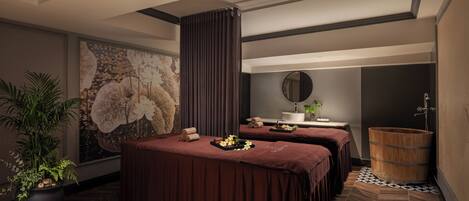 A spa room with two massage tables, a large wall mural, a potted plant, a wooden barrel, and a mirror.