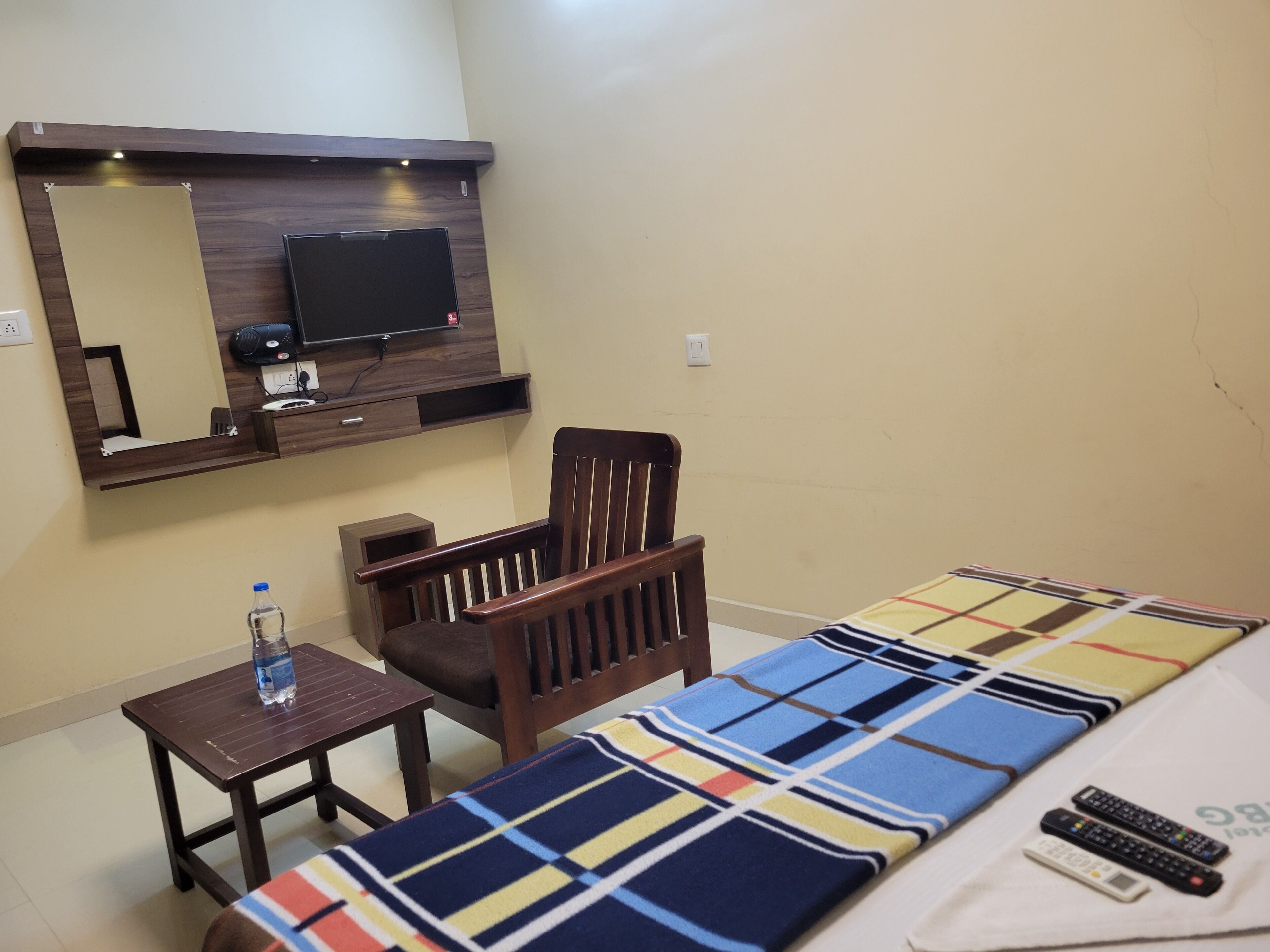 Una habitación de hotel con una cama, una silla de madera, una mesita con una botella de agua, un televisor fijado en un estante de madera y una manta estampada.