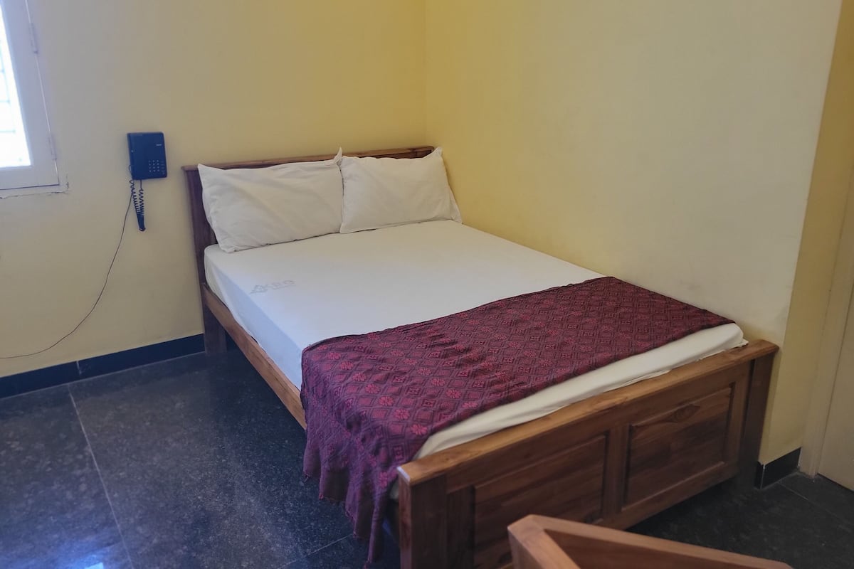 Una cama individual con estructura de madera, ropa de cama blanca y una manta estampada. Un teléfono está fijado a la pared a la izquierda.